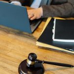 Vue d'en haut d'un avocat anonyme en tenue formelle tapant sur un ordinateur portable assis à une table en bois avec une pile de documents et un marteau